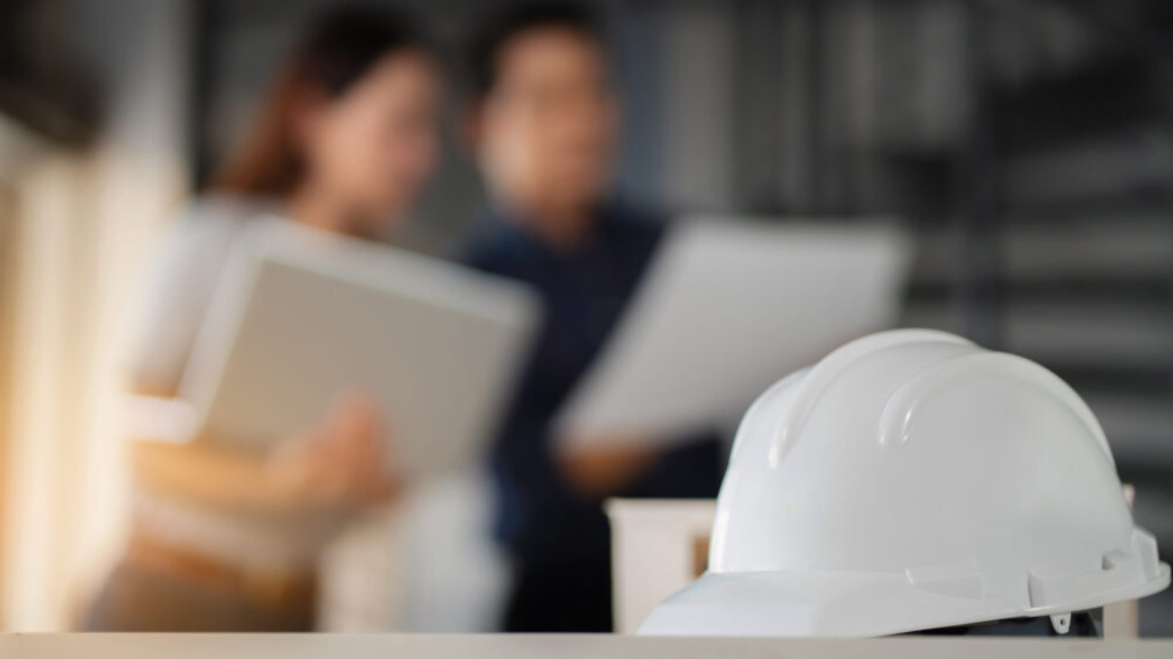 White safety helmet on a desk with two construction professionals reviewing documents in the background, representing the role of financial software for construction in Ireland.