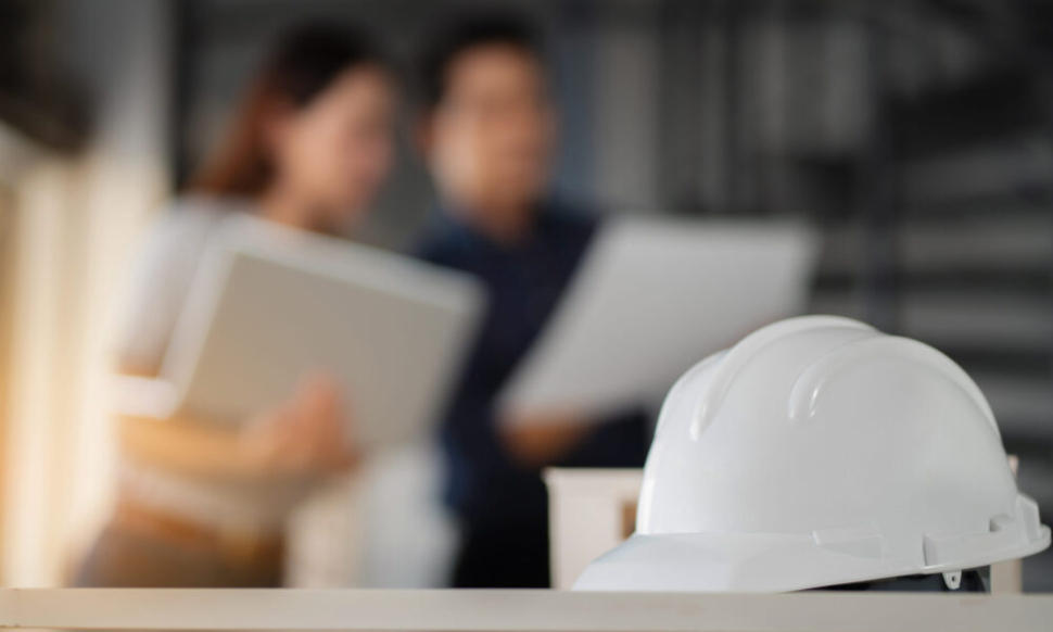 White safety helmet on a desk with two construction professionals reviewing documents in the background, representing the role of financial software for construction in Ireland.