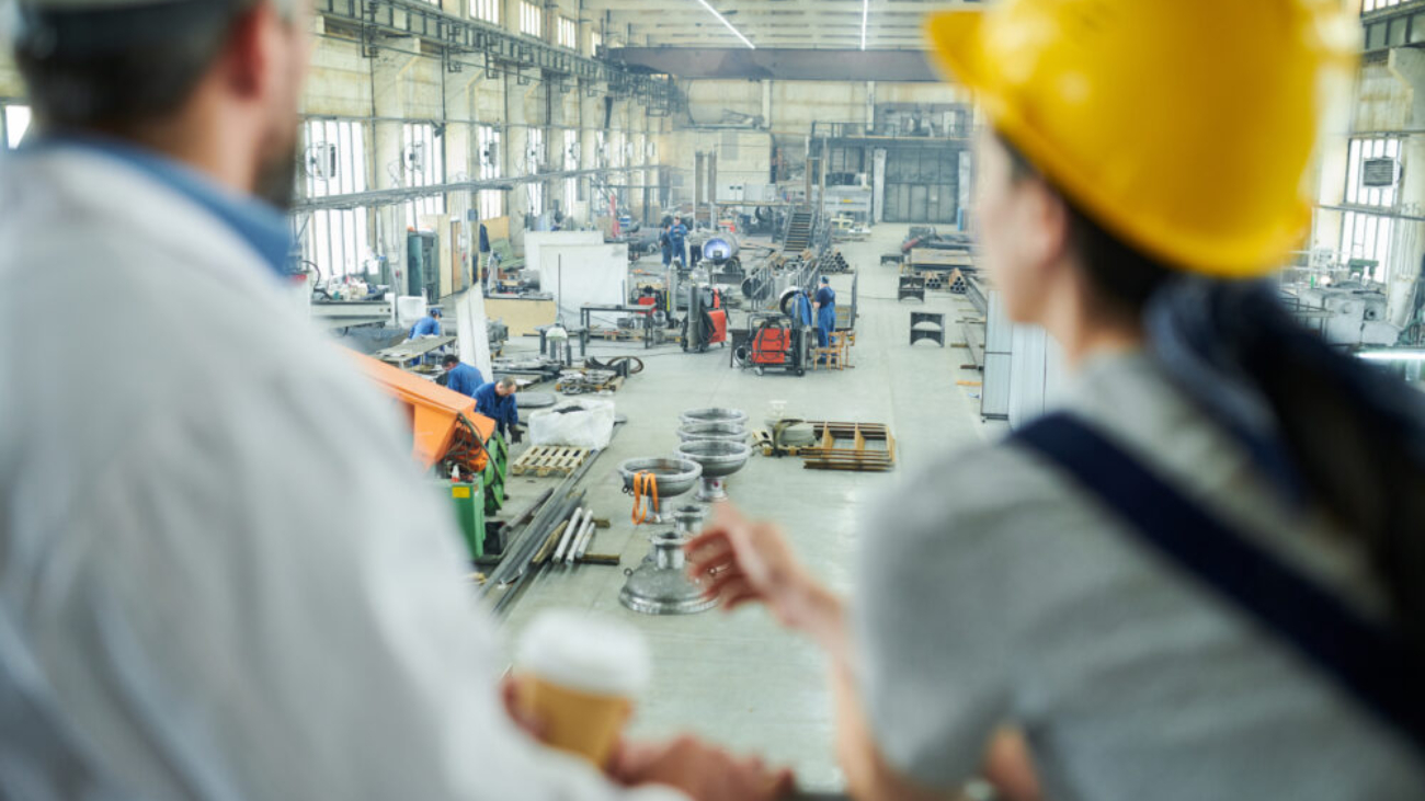 Engineers overlooking a busy factory floor with workers and machinery, highlighting the role of manufacturing software systems in managing production operations.