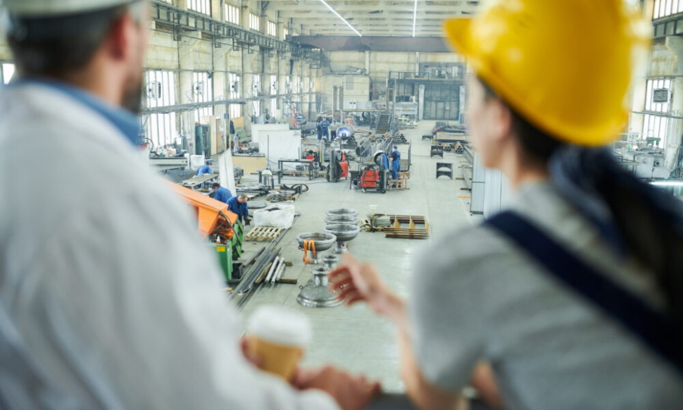 Engineers overlooking a busy factory floor with workers and machinery, highlighting the role of manufacturing software systems in managing production operations.